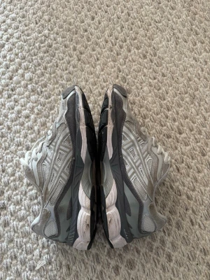 Gråa Asics sneakers med meshdetaljer - Säljer ett par gråa Asics sneakers med vita och mörkgrå detaljer. Skorna har klassisk mesh och syntetmaterial i ovandelen, samt Asics-logga på sidan. Sulan är vit med inslag av svart och mörkblått. Perfekta för dig som gillar sportig stil.