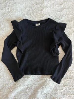 Svart ribbad topp med volang  - Söt tröja i svart ribb material med fina volanger vid axlarna. Mjukt och skönt material i mycket fint skick men som blivit för liten för mig. Från Zara i storlek 134cm.