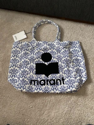 Isabel Marant Tote Bag tyg - Snygg sommarväska från Isabel Marant i vitt tyg med blått, dekorativt mönster. Stor svart logga och text framtill. Väskan har två handtag och är rymlig, perfekt för att bära med sig allt man behöver. Materialet ser ut att vara kraftig bomullscanvas.