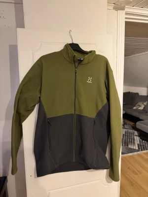 Haglöfs kofta  - Säljer en grön och mörkgrå fleecejacka från Haglöfs med hel dragkedja framtill och två sidofickor med dragkedja. Jackan har hög krage och är tillverkad i mjukt fleecematerial, perfekt för lager-på-lager och outdoor.