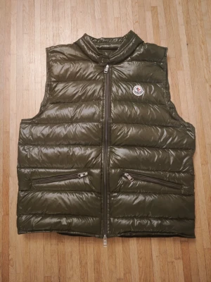 Moncler Gui Down Väst. Strl 3. Nyskick - Moncler Gui Down Vest. Använd två gånger och säljes på grund av att den är för stor för mig. Passar S och M. Västen är i nyskick som du kan se på bilderna. Sden är grön med glansig finish och ståkrage. Västen har två dragkedjeförsedda fickor framtill och klassisk Moncler-logga på bröstet. Perfekt för lager-på-lager och håller dig varm under kyliga dagar.