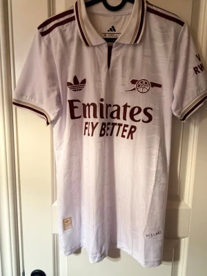 Arsenal x Adidas retrotröja vit - Vit Arsenal retrotröja från Adidas med vinröda detaljer och broderade loggor. Klassisk krage med ränder, tryckt sponsortext och klubbmärke på bröstet. Tillverkad i lätt och ventilerande material, perfekt för fotboll eller träning.