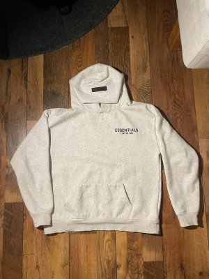 Grå Essentials Fear of God hoodie - Snygg grå hoodie från Essentials Fear of God med svart logga på bröstet och stor text på ryggen. Använd få gånger så den är som ny. Kommer med taggen och påse. Kvitto finns även, ny pris är 1500. Klassisk oversized passform, stor huva och magficka. Pris kan diskuteras. Vid funderingar är det bara att fråga!