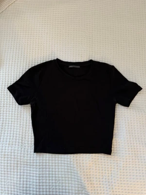 Svart croppad topp från Zara - En enkel och stilren svart croppad topp från Zara. Toppen har rund halsringning och korta ärmar, perfekt att matcha med höga jeans eller kjol. Tillverkad i mjuk bomull för en bekväm känsla.