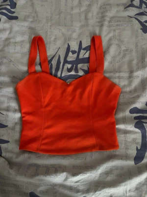 Orange croppad linnetopp med dragkedja - Snygg orange croppad linnetopp med breda axelband och hjärtformad urringning. Toppen har en synlig dragkedja i ryggen och är gjord i ett stretchigt material som sitter tight. Väl använd men bra skick.