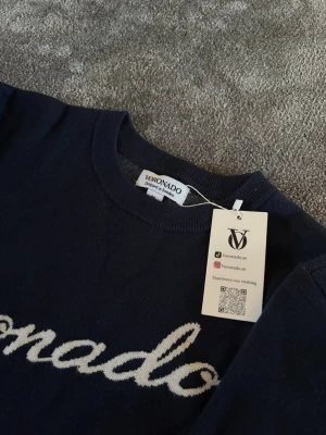 Mörkblå tröja från Voronado - Mörkblå tröja från Voronado med vit broderad logga över bröstet och på ärmen. Tröjan har rund halsringning och är tillverkad i ett mjukt material som känns skönt mot huden. Perfekt för en stilren och avslappnad look.