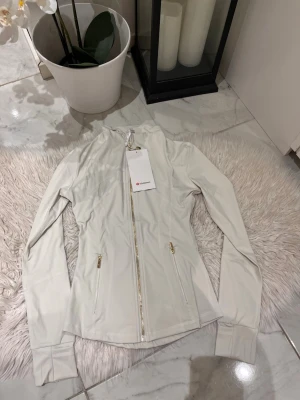 Vit zip hoodie från Lululemon - Snygg vit zip hoodie från Lululemon med guldfärgade dragkedjor och figurnära passform. Tillverkad i ett stretchigt funktionsmaterial som passar perfekt till träning eller chill. Två fickor med dragkedja och hög krage för extra stilpoäng.