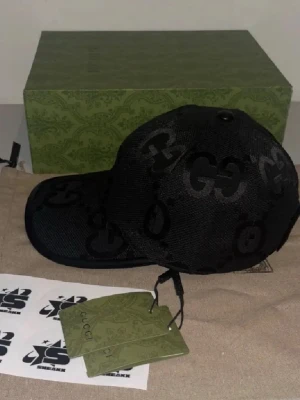 Svart keps från Gucci med GG-mönster - Snygg svart keps från Gucci med klassiskt GG-mönster över hela kepsen. Kepsen har böjd skärm och justerbar baksida. Tillverkad i mesh-liknande material som ger en modern och luftig känsla. Perfekt för dig som vill ha en exklusiv och trendig accessoar.