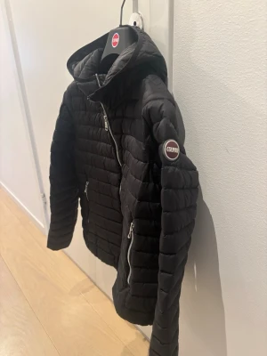 Svart jacka från Colmar - Svart dunjacka från Colmar med huva och två dragkedjeförsedda fickor framtill. Jackan har Colmar-logga på ärmen. Perfekt för våren och enkel att matcha med olika outfits. Storlek 160 (14år)