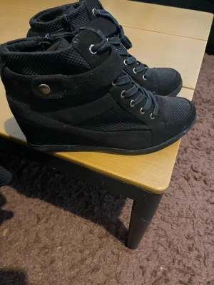 Svarta wedge sneakers med meshdetaljer - Säljer ett par svarta sneakers med kilklack och meshdetaljer. Skorna har snörning, kardborreband och metallnitar vid sidan en sportig men ändå edgy look. Perfekta för dig som vill ha lite extra höjd i vardagen.