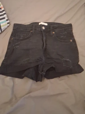 Svarta denimshorts med hög midja - Snygga svarta jeansshorts med hög midja och råa kanter. Klassisk femficksmodell med knapp och dragkedja framtill. Slitna detaljer ger en cool och avslappnad look. Perfekta till sommaren och lätta att matcha med olika toppar.