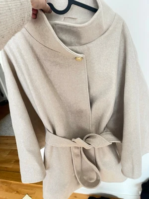 Beige kappa med knytskärp - Stilren beige kappa med hög krage och knytskärp i midjan. Kapp hiding en dold knäppning med en dekorativ guldfärgad knapp upptill. Materialet ser ut att vara mjukt och lite tjockare, perfekt för kyligare dagar. Rak passform och vida ärmar ger en modern, avslappnad look.