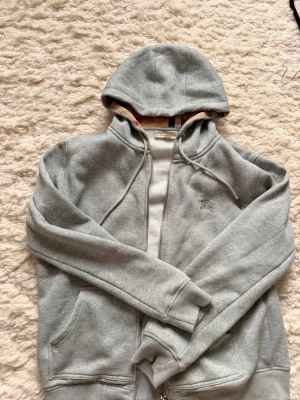 Grå zip hoodie från Burberry - Säljer en stilren grå zip hoodie från Burberry med klassiska snören i huvan och fickor framtill. Hoodien har dragkedja och den ikoniska Burberry-randen syns på insidan av huvan. 