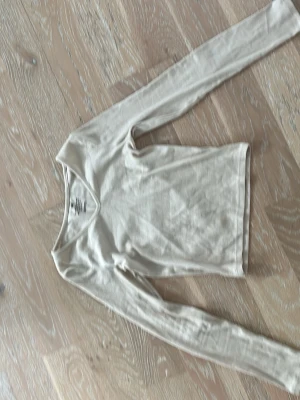 Beige ribbad långärmad topp H&M - Säljer en beige ribbad långärmad topp från H&M med v-ringning. Toppen är i mjuk bomull och har en slim passform som sitter snyggt på kroppen. Perfekt att styla med jeans eller kjol för en enkel och clean look.