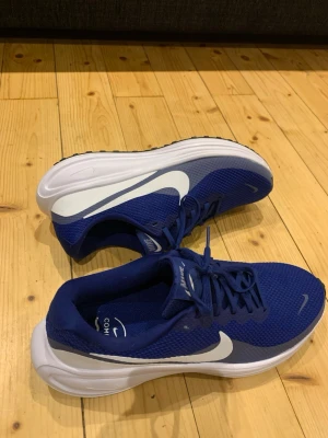 Blå Nike sneakers med vit sula - Snygga blå sneakers från Nike med vit sula och den klassiska vita swoosh-loggan på sidan. Ovandelen är i meshmaterial för bra andningsförmåga och skon har svarta detaljer. Perfekta för dig som vill ha både stil och komfort. Det är både en bra vardagsko men framförallt löparsko. Otroligt sköna!