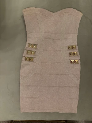 Champagnefärgad axelbandslös klänning - Axelbandslös kort klänning i champagnefärgad glittrig textil med markerade sömmar och dekorativa guldfärgade nitar framtill. Klänningen har figurnära passform och dragkedja bak. Perfekt för dig som vill sticka ut.