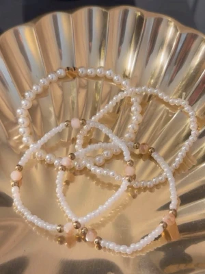 Pärlarmband med guldiga detaljer - Säljer ett set med fyra elastiska armband i vitt och guld. Armbanden är gjorda av små vita pärlor, några med inslag av ljusrosa pärlor och guldfärgade detaljer. Ett av armbanden har en liten guldfärgad hjärtberlock. Perfekt för att ge din outfit en söt och trendig touch.🥹🧸🩷