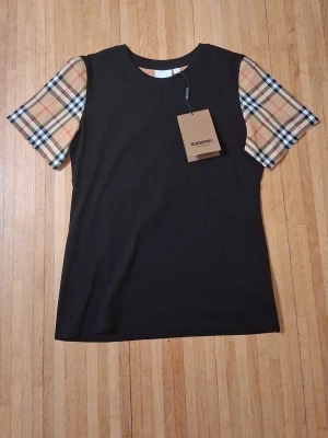 Burberry svart t-shirt med rutiga ärmar - Säljer en svart t-shirt från Burberry med klassiska rutiga korta ärmar i beige, svart och röd. T-shirten har en enkel rund halsringning och är tillverkad i mjuk bomull. Perfekt för dig som vill ha en stilren look med en touch av lyx. Passar också M