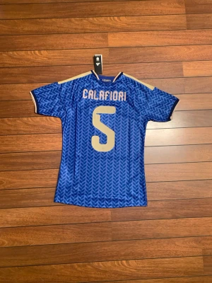 Italien matchtröja Calafiori #5 blå - Snygg blå Italien matchtröja från Adidas med Calafiori och nummer 5 på ryggen. Tröjan har gulddetaljer, tre ränder på axlarna och Italiens landslagsemblem på bröstet. Tillverkad i lätt och ventilerande material, perfekt för fotboll eller träning.