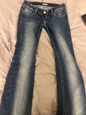 Blå lowwaist bootcut jeans med broderade fickor - Säljer ett par blå väldigt lowwaist bootcut jeans med snygg slitning framtill och broderade bakfickor med lock och knapp. Jeansen har klassisk femficksmodell och är gjorda i denim med stretch. Perfekta för dig som gillar detaljer och en avslappnad look. Byxorna är från Ginatricot och är i fint skick och ej användts många gånger, säljer pga att de är för små. 
