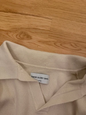 Beige tröja från These Glory Days - Snygg beige tröja från These Glory Days med krage och v-ringning. Tillverkad i mjukt stickat material som ger en clean och stilren look. Perfekt för dig som gillar minimalistisk stil och vill ha något enkelt men ändå trendigt.