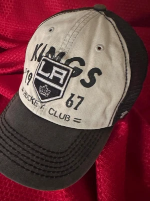 LA Kings svart och vit keps - Snygg LA Kings keps i svart och vitt med broderad logga och text framtill. Kepsen har böjd skärm, mesh på sidorna och justerbar snapback baktill. Perfekt för dig som gillar hockey och vill ha en sportig stil.