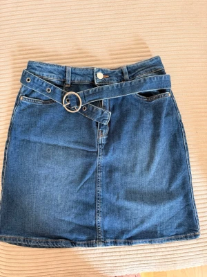 Blå jeanskjol med bälte - Trendig blå jeanskjol med tillhörande denim-bälte och silverfärgat spänne. Kjolen har klassisk femficksdesign och kort längd, perfekt för en avslappnad och cool stil. Passar till många olika toppar och sneakers.