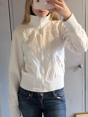 Vit vindjacka med hög krage - Snygg vit vindjacka med hög krage och dragkedja framtill. Jackan har ribbade muddar vid ärmslut och midja samt två fickor med dragkedja. Perfekt för blåsiga dagar och enkel att matcha till olika outfits.