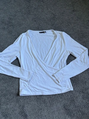 Vit omlottopp med lång ärm - Snygg vit omlottopp med lång ärm och djup v-ringning. Tunn och mjuk i materialet, perfekt för lager på lager eller som den är. Enkel och stilren design som passar till mycket.