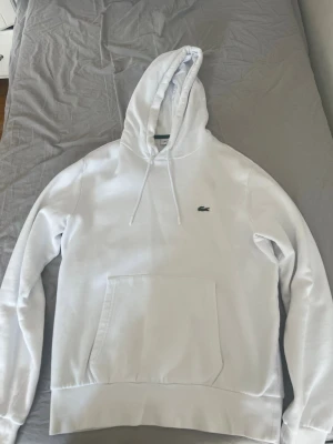 Vit Lacoste hoodie - vit hoodie från Lacoste med klassisk krokodillogga på bröstet. Hoodien har huva med dragsko, stor magficka och ribbade muddar vid ärmslut och midja. Perfekt för en clean och stilren look. Materialet känns mjukt och skönt mot huden. 