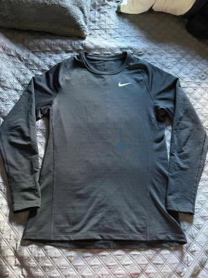 Svart långärmad träningströja Nike - Svart långärmad träningströja från Nike med Dri-FIT-teknologi och Nike Pro-detalj. Tröjan har rund halsringning och en vit Nike-logga på bröstet. Perfekt för träning och sportaktiviteter.
