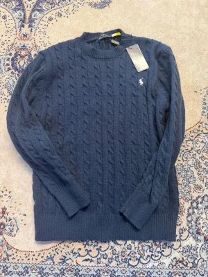Mörkblå kabelstickad polo tröja  - Mörkblå kabelstickad tröja från U.S. Polo Assn. med rund halsringning och diskret logga broderad på bröstet. Tröjan har långa ärmar och ribbade muddar vid ärmslut och nederkant. Perfekt för lager på lager och klassisk stil.