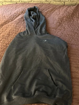 Mörkgrå hoodie från Gymshark - Snygg mörkgrå hoodie från Gymshark med klassisk huva och magficka. Diskret Gymshark-logga på bröstet. Perfekt för chill eller träning. Mjuk bomullskänsla och ribbade muddar vid ärmslut och nederkant.