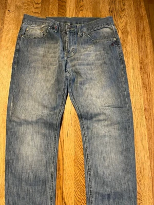 Schyssta dondup jeans med slitningar  - Säljer ett par klassiska Dondups blå jeans med raka ben och snygga slitningar. Jeansen har fem fickor och stängs med dragkedja och knapp. Perfekta för en avslappnad stil och passar till det mesta. Materialet är denim i bomull.