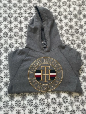 Grå hoodie från Tommy Hilfiger - Snygg grå hoodie från Tommy Hilfiger med stor guldbroderad logga och text på! Den är använd men några få gånger men helt ny! Inga fläck eller slitage, priset går att diskutera! 