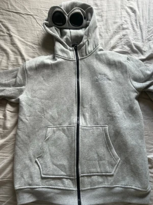 Grå hoodie med goggles från C.P. Company - Säljer en grå hoodie från C.P. Company med hel dragkedja, känguruficka och ikoniska goggles i huvan. Broderad logga på bröstet. Mjuk bomullsblandning och ribbade muddar. Perfekt för dig som gillar streetwear och unika detaljer. Har lossnat en knapp från den annars inga defekter.