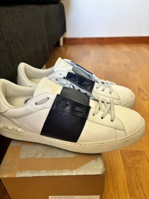 Valentino Garavani vita sneakers - Snygga vita sneakers från Valentino Garavani med bred svart läderdetalj över snörningen. Klassisk låg modell med diskreta nitar på hälen och vita skosnören. Tillverkade i läder för en lyxig känsla och stilren look.