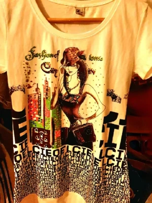 Vit t-shirt med Rhinestone - Passar S, M och förmodligen L. Cool vit t-shirt med färgglatt tryck av en tecknad tjej, stadssilhuett och text. T-shirten har glittriga paljettdetaljer och korta ärmar. Perfekt för dig som gillar unika och trendiga motiv.
