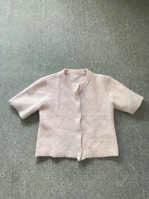 Ljusrosa stickad kofta med kort ärm - Söt ljusrosa stickad kofta med korta ärmar och rund halsringning. Koftan har knäppning framtill med matchande knappar och är gjord i ett mjukt, ribbstickat material. Perfekt att slänga över en topp för en chill look. Helt ny.
