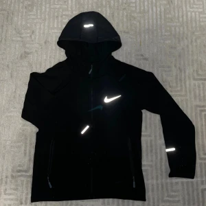 Svart vindjacka med huva. Nike 160-170cm - Svart vindjacka med huva och reflekterande detaljer, perfekt för träning utomhus. Jackan har dragkedja framtill, två sidofickor med dragkedja och en stor vit logga på bröstet. Materialet är lätt och syntetiskt, vilket gör den smidig att bära.