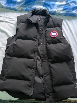 Canada Goose - Snygg svart dunväst från Canada Goose med klassisk logga på bröstet. Västen har hög krage, dragkedja och tryckknappar framtill. Perfekt för lager-på-lager och håller dig varm med sin vadderade design.