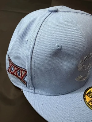 Ljusblå Supreme x New Era keps - Snygg ljusblå keps från Supreme i samarbete med New Era. Kepsen har broderade detaljer, bland annat Supreme-loggan i rött och vitt på sidan samt en glittrig S på framsidan. Klassisk form med böjd skärm och ventilation på ovansidan.