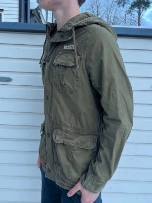 Olivgrön vindjacka från Hollister - Snygg olivgrön vindjacka från Hollister med huva och flera praktiska fickor framtill. Jackan har dragkedja och knappar, samt en avslappnad passform. Perfekt för blåsiga dagar och enkel att matcha med jeans eller chinos.