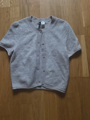 Beige kortärmad kofta från H&M - Kortärmad beige kofta från H&M med rund halsringning och knappar framtill. Koftan är stickad i mjukt material och har ribbade kanter vid hals, ärmslut och knappslå. Perfekt att slänga över en topp för en chill look.