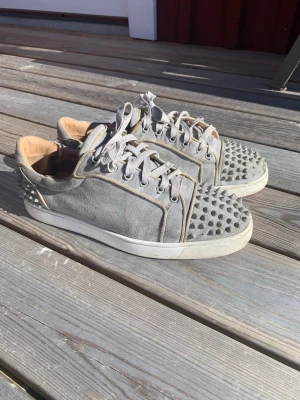 Gråa sneakers med nitar från Louboutin - Unika gråa sneakers i mocka från Christian Louboutin med coola nitar på tån och hälen. Perfekta för dig som vill sticka ut med din stil och gillar exklusiva detaljer. Negativit att vissa nitar är borta. 