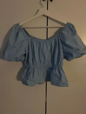 Ljusblå offshoulder blus med puffärm - Supersöt ljusblå blus med offshoulder och puffiga korta ärmar. Smockad i midjan för en snygg passform och volang nertill. Perfekt för en trendig och somrig look.