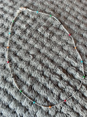 Tunt halsband med färgglada pärlor - Ett tunt och stilrent halsband med små pärlor i olika färger som blått, grönt, rött och orange. Kedjan är silverfärgad och halsbandet har en minimalistisk design som ger en lekfull touch till din outfit.