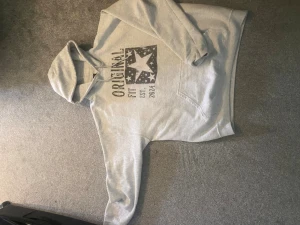 Grå hoodie med tryck och huva - Snygg grå hoodie med stor stjärna och texten 'ORIGINAL FIT EST. 2024' på bröstet. Klassisk modell med huva och magficka. Perfekt för en avslappnad och trendig stil. Perfekt för 181 till 185 