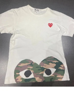 Vit Comme des Garçons PLAY t-shirt - Vit t-shirt från Comme des Garçons PLAY med ikoniskt rött hjärta på bröstet och stort camouflagemönstrat ögonmotiv längst ner. Klassisk passform och rund hals. Tillverkad i mjuk bomull som känns skön mot huden.