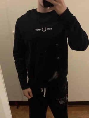 Svart sweatshirt från Fred Perry - Svart sweatshirt från Fred Perry med vit broderad logga och text på bröstet. Klassisk rund halsringning och ribbade muddar vid ärmslut och nederkant. Perfekt för en stilren och avslappnad look.